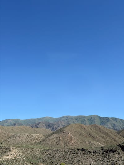 The image depicts a vast, arid mountainous landscape under a clear, bright blue sky. The foreground is dominated by rolling hills covered in sparse, dry vegetation and rocks, with textures ranging from coarse gravel to larger stones. Some areas show faint green patches, suggesting scattered plant life. As the eye moves towards the background, the terrain becomes more rugged, with distinct mountain ranges layered upon one another. These further mountains are a deeper green, their slopes carved with numerous ravines and gullies, indicating erosion. The furthest range appears softer and more distant, almost blending into the horizon. There are no visible people, structures, or immediate signs of human activity. The environment is natural, characterized by its rugged terrain, limited vegetation, and clear atmospheric conditions. The scene is consistent with a high-altitude, dry climate. The location in Kok Moynok Eki, Kyrgyzstan, suggests a remote, natural area.