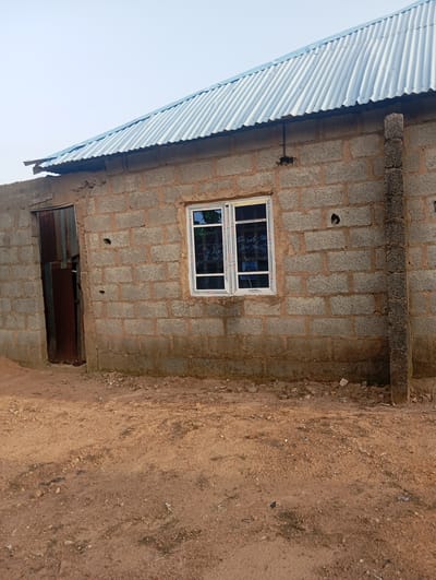 The image shows a close-up, eye-level, medium shot of the exterior of a building under construction in Karsana 2, Nigeria. The building is constructed from grey concrete blocks with visible mortar. A corrugated metal roof, light blue in color, is installed. On the left side of the building, there is a partially open metal door with visible rust. In the center of the building, there is a modern white-framed window with clear glass, reflecting the sky and some trees. The ground in front of the building is a dirt surface with scattered small stones and debris. A vertical concrete pillar is positioned on the right side of the building, near the edge of the frame. The sky is clear and light-colored, suggesting daytime. There are no people or animals visible in the image.