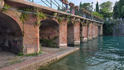 The image shows a brick bridge with multiple arches crossing a body of turquoise water. The bridge is covered in greenery and has flower boxes lining its upper railing, where several people are visible walking. The water has ripples and reflections of the bridge and the surrounding trees. The area under the arches appears to be paved with cobblestones, and graffiti is visible on the brick walls. The scene is set in Peschiera del Garda, Italy, with lush green trees and a stone embankment visible on the right side of the frame.