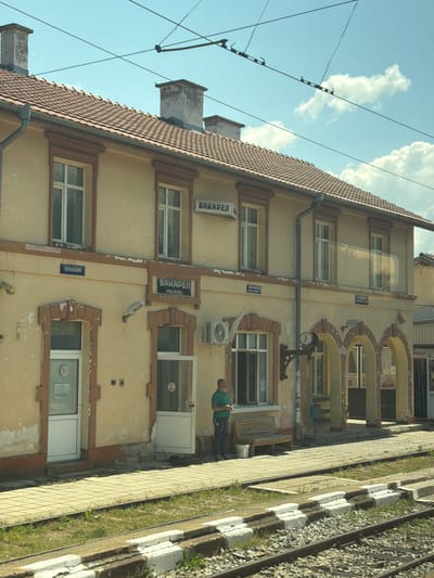 The image shows the exterior of a building, likely a train station, with visible signage in Bulgarian. The building has a tiled roof and a pale yellow facade. There are several windows and arched entrances. In front of the building, there is a paved platform with train tracks and gravel on the side. A man in a green shirt and dark pants is standing near the building, facing left. To his left, there is a wooden bench with a white bucket underneath. The sky is blue with some clouds. The overall impression is of an outdoor scene in a rural or small-town setting. Based on the signage, the location is identified as the train station in Baliovtsi, Bulgaria.