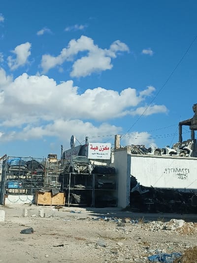 The image depicts an outdoor scene in Al-Qadam Municipality, Syria, showing a commercial area primarily associated with vehicle salvage or spare parts. The foreground consists of an unpaved, uneven dirt and gravel surface littered with debris, including concrete fragments, plastic bags (one black, one blue), and general refuse.

To the left, a structure resembling an automotive parts yard is visible. It features tall, multi-level metal shelving units stacked with numerous vehicle components, including tires, engine parts, body panels, and what appears to be a stripped car chassis positioned atop a shelf. Two wooden crates are on the ground in front of these shelves. A white banner with red Arabic text reading "مخزن هبة" (Makhan Hiba - Hiba Store/Warehouse) is affixed to the building behind the shelves. A circular logo, possibly a stylized "VW" for Volkswagen, is partially visible above this banner.

To the right, a white wall-like structure extends into the frame. Spray-painted on this wall are Arabic numbers ".٩٦٣٨٨٨٢٢١" (likely a phone number), followed by "للتعهد" (for contracting), and "أبو خالد" (Abu Khaled). Behind and above this wall, additional vehicle wreckage and components, including several white car body frames, are stacked. The upper portion of the structure to the far right appears to be significantly damaged or incomplete, with exposed rebar and broken concrete elements.

The background is dominated by a clear blue sky dotted with scattered white cumulus clouds. A thin, dark power or communication line stretches diagonally from the top left towards the mid-right of the image. No individuals are present in the scene, and no overt actions or interactions are depicted. The overall impression is one of a functional but somewhat dilapidated industrial or commercial zone specializing in vehicle parts, set against a bright, sunny day.