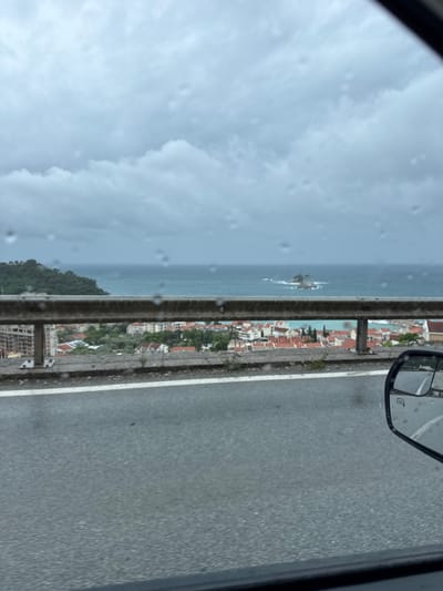 The image shows a view from a vehicle traveling along a road adjacent to the Adriatic Sea in Medinski Krš, Montenegro. The sea, with small rocky islands, is visible on the left, and the town's buildings, distinguished by red-tiled roofs and dense greenery, are situated on the coast. A grey metal guardrail runs parallel to the road, separating it from the seaside view. The road surface is asphalt, marked with a white lane divider. The vehicle's side mirror is visible in the upper right corner of the image, and the front windshield partially obscures the left side of the frame, exhibiting raindrops. The sky is overcast.