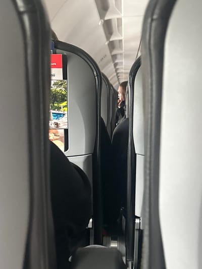 The scene is an aircraft interior, contextually located in the city of Fischamend, Austria, although no specific geographical landmarks are visible within the cabin itself. The image is captured from an aisle perspective, looking forward or backward between two rows of seats.
In the immediate foreground, the view is partially obscured by the backs of two light-grey aircraft seats, framing the central focus. The seat on the left features a seat-back pocket containing a partially visible magazine or pamphlet. Red text "CAFE" and "SÜ" are discernible on the upper portion of the visible publication. Below this, an image depicting a green lawn adjacent to a swimming pool with lounge chairs is visible, followed by a partial view of what appears to be food at the bottom. The upper legs and lap of a person, dressed in black clothing, are visible beneath this seat. Similarly, the upper legs and lap of another person in black clothing are visible beneath the seat on the right.
Further down the aisle, in the mid-background, a passenger with a short haircut or shaved head is visible in profile. This individual appears to be holding a phone or object to their ear and is wearing dark clothing. The cabin ceiling is white with visible overhead panels and vents. The interior is artificially lit.