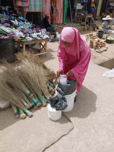 This image captures an outdoor market scene in Kadoma, Zimbabwe. In the foreground, a woman dressed in a bright pink full-body covering and headscarf bends over, handling items within a white plastic bucket containing a black plastic bag. Another smaller black plastic bag rests on a second, smaller white bucket beside it. To her left, a large pile of natural fiber brooms, each bound with green or light-colored tape at the handle end, lies on the cracked concrete ground. The background reveals additional market activity: to the left, a stall displays shoes and other merchandise on a wooden table, with an individual seated behind it. To the right, piles of produce, resembling root vegetables, are arranged on the ground, and other vendors and shoppers are visible further back, including one wearing a straw hat. The ground transitions from concrete in the foreground to packed earth or sand in the background.