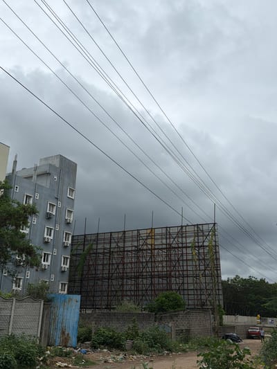 The image captures an overcast day in Shamshabad, India, with a focus on urban infrastructure. In the foreground, a dirt path is littered with various types of debris, including plastic bags and wrappers, indicating a disregard for waste disposal. To the left, a low, grey concrete wall with an ornate pattern runs along the path. Adjacent to it is a tall, rusted, blue metal shed. Behind these elements, a multi-story building stands, its grey facade punctuated by rows of white-framed windows and air conditioning units. To the right of the building, a large, skeletal billboard structure, made of interconnected metal beams, dominates the mid-ground. Green vines are creeping up the structure. Further to the right, a red car is parked on a dirt surface, with two individuals standing nearby. A wider concrete barrier encloses the area in the distance, with lush green trees forming a backdrop. Overhead, a network of dark power lines crisscrosses the grey, cloudy sky. The overall scene suggests an area undergoing development or experiencing neglect, with a juxtaposition of modern construction and the harsh reality of urban waste.
