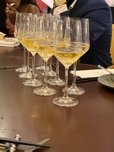 A long shot from eye level shows a line of seven champagne flutes filled with a bubbly, golden-colored liquid, arranged in a row on a dark brown table. The glasses are clear and have thin stems, and they are spaced evenly apart. The flutes are oriented diagonally from the viewer's perspective, with the nearest glass prominently displayed. The liquid in the glasses exhibits numerous small bubbles, indicating it is carbonated. The background is largely out of focus. On the left side of the frame, a person wearing a pink top is partially visible, holding a small object in their hand near their mouth. To the right of the glasses, a person in a dark blue jacket is visible, wearing a gold wristwatch. A white napkin and a decorative silver dish are also on the table. The setting appears to be indoors, possibly a restaurant or event space. The location in Xuanwu District, China, is not evident from the image itself.