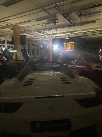 The image shows a collection of high-end cars in what appears to be a garage or showroom in Wetzikon (ZH), Switzerland. Dominating the foreground is a white Ferrari with its door open. A red car is visible behind it. The word "Ferrari" is visible on the body of the white car, along with the "VETTIGER AUTOMOBILE" logo. The lighting is predominantly artificial, coming from the building's structural elements. Fluorescent lighting structures are visible in the ceiling. The environment is modern and appears to be well-maintained. Additional elements include signs with glowing lighting within the backdrop of the garage.