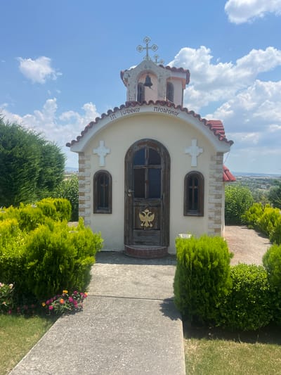 The image shows a small, cream-colored church with a red-tiled roof, set against a bright blue sky with fluffy white clouds. The church has a distinctive arched doorway with dark wooden doors, which are decorated with a golden eagle emblem. Above the door, there is a small, arched window. To the left of the door, there is a white cross with a slightly ornate design, and a bell is visible in the belfry. The exterior of the church is adorned with decorative stone elements along the top of the facade and a curved, tiled roofline.

The church is surrounded by lush green bushes and some flowering plants, with a concrete path leading up to it. In the background, a hilly landscape with scattered greenery can be seen, suggesting a rural or semi-rural setting. The time of day appears to be daytime, with bright sunlight casting shadows, and the weather seems pleasant.

Visible text on the church, written in Greek letters, appears to say "Ι. Ν. ΤΟΝΝΕΟΥ ΠΡΟΔΡΟΜΟΥ" which likely translates to "Holy Church of John the Baptist". The overall impression is one of a serene and picturesque religious building in a natural setting. The image was taken in Kardia, Greece.
