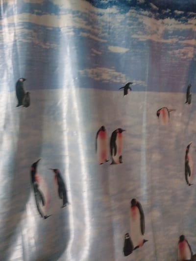 The image depicts a shower curtain with a pattern of penguins. There are various penguins of different sizes and poses, some standing, some appearing to waddle, and one in mid-air. The background of the curtain is a light blue with white streaks, resembling clouds or sky. The image is taken indoors, likely in a bathroom, with a slight sheen reflecting off the curtain. There is no indication of the city of Ananyevo, Kyrgyzstan, in the image.