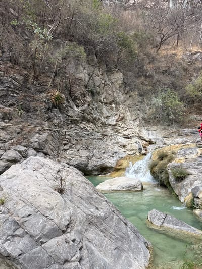 The image depicts a natural canyon landscape in San Miguel Atlapulco, Mexico, characterized by a stream, waterfall, and rocky terrain under an overcast sky. In the foreground, large, grey, angular rock formations with visible striations dominate the lower left. A clear, greenish-blue stream flows from right to left, encompassing these rocks and several submerged boulders. In the midground, a small waterfall cascades approximately one meter into the stream from a higher, yellowish-tan rock formation, indicating mineral deposits. A horizontal line, resembling a cable or rope, spans across the scene, affixed to the steep rock face above the water. A person, partially visible on the far right, wears a bright red top and appears to be standing on the stream bank. The background consists of a steep, rugged cliff face of grey rock, densely covered with sparse, dry, deciduous vegetation and several agave-like succulent plants. The overall environment suggests a dry or semi-arid climate.