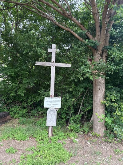 This media file shows a wooden cross and a stone plaque in a natural outdoor setting. The cross is weathered and appears to be made of rough-hewn wood. Attached to the cross is a white sign with text written in Cyrillic script. To the right of the cross, a stone plaque is set into the ground, featuring an embossed image and more Cyrillic text. The background is filled with lush green foliage, trees with thick trunks and branches, and some dry earth and grass. The overall impression is of a solemn or commemorative site, possibly a memorial or the beginning of a construction project in a rural area. The text on the sign indicates that the location was consecrated on June 18, 2008, for the construction of a church chapel in honor of Saint Sergius of Radonezh. This suggests the image captures a significant moment in the establishment of a religious building. The lighting and foliage suggest it is daytime, likely during the warmer months. The scene appears peaceful and undisturbed, with no people visible.