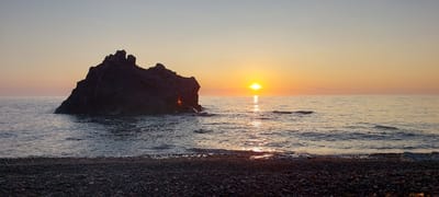 The image depicts a tranquil seaside scene at dusk, likely in the vicinity of Aleksandrovsk-Sakhalinskiy, Russia, given the context provided. The foreground is a beach covered in dark, multi-colored pebbles. The sea gently laps at the shore, with small waves creating a shimmering effect on the water's surface. To the left, a large, dark rock formation juts out from the water, silhouetted against the warm hues of the sunset. The sun, a bright, intense orb, is setting on the horizon, casting a golden and orange glow across the sky and reflecting a path of light on the calm sea. The sky transitions from a pale blue at the top to soft oranges and pinks near the horizon. A few small birds can be seen in the distance, adding a sense of scale and life to the vast seascape. The overall atmosphere is serene and natural. There are no people or visible man-made structures in the frame.