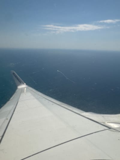 The image shows a view from an airplane window looking down at the sea and sky. The perspective is from high altitude, with a plane wing visible in the foreground, stretching diagonally across the right side of the frame. The wing is white and shows some light and shadow patterns.
In the distance, the sea appears as a dark, deep blue expanse. A faint contrail is visible in the sky above the sea, a thin white line stretching horizontally. The sky on the left side of the image is a lighter blue, with some wispy white clouds. The overall impression is of a clear day, likely sometime during daylight hours, given the brightness.
There are some scratches and smudges on the airplane window, which are visible throughout the image, creating a slightly hazy effect.
A notable detail is the text visible on the tip of the airplane wing. It appears to be a logo or text, possibly identifying the airline. The visible part of the text seems to be "RYANAIR".
The scene captures the typical view one might experience during an airplane journey. There are no people visible in the image, and the focus is on the aerial landscape and the aircraft's wing. The image is oriented vertically, suggesting it was taken with the camera held in a portrait orientation.