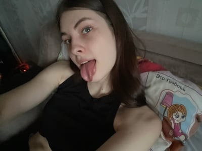 A person is lying on a bed with a pillow. The person has long brown hair, fair skin, and is sticking their tongue out. The person is wearing a black top. The pillow has a cartoon image on it with text in Russian. The environment appears to be a bedroom with a bed and other bedding materials. There is no information available to suggest this photo was taken in Shcheglovo, Russia.