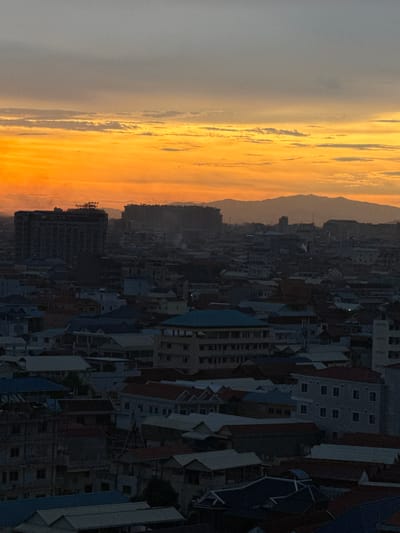 This is a high-angle, medium shot of an urban landscape at dusk, likely in Khan Mean Chey, Cambodia. The sky is dominated by a dramatic sunset with vibrant orange and yellow hues blending into a pale grey at the top. Wispy clouds are scattered across the horizon. Below the sky, a dense collection of buildings fills the frame, varying in size and architectural style.

The buildings are mostly residential or low-rise commercial structures, with a variety of roof colors including red, grey, and blue. Some taller, darker buildings, possibly under construction or older structures, are visible in the mid-ground, silhouetted against the bright sky. The overall impression is one of a developing urban area.

There are no visible people or specific actions depicted in the scene. The environment is characterized by the dense urban sprawl and the atmospheric lighting of sunset. The image focuses on the cityscape and the sky, creating a mood of quiet observation.