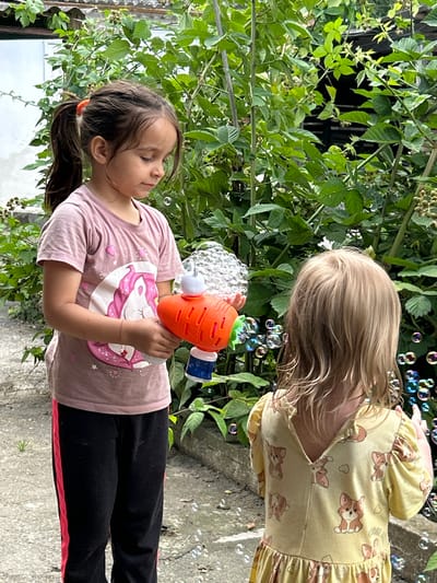 Two young girls are playing with a bubble gun in a garden. The girl in the foreground, with dark hair tied in a ponytail, wears a pink t-shirt with a unicorn design and black pants with a pink stripe. She is holding an orange carrot-shaped bubble gun, from which bubbles are emerging. The girl in the background, with blonde hair, wears a yellow dress with a pattern of corgi dogs. Bubbles are floating around both girls. The setting appears to be a backyard with green foliage, including what looks like berry bushes, and a concrete ground. The lighting suggests it is daytime, and the weather appears to be pleasant, conducive to outdoor play. There is no visible text in the image.