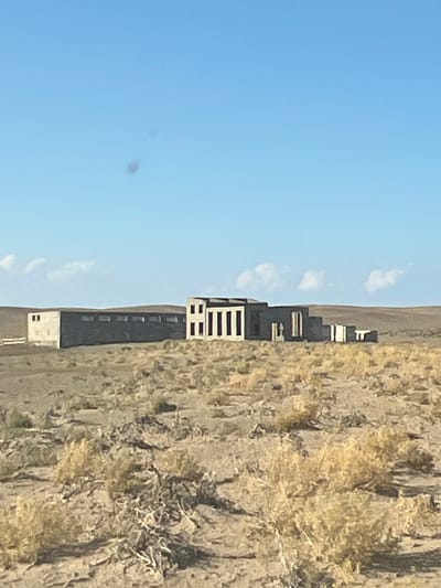 The image shows a complex of concrete structures in a desolate, arid landscape. The structures appear to be abandoned and are partially integrated into the undulating terrain. The sky is a clear blue with a few scattered clouds. The foreground is dominated by dry, sparse vegetation, characteristic of a desert environment. The structures themselves are geometric and functional in design, suggesting they may have been industrial or institutional buildings. Their weathered appearance and the surrounding emptiness evoke a sense of isolation and decay.