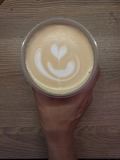 A clear plastic cup of coffee with latte art in the shape of a heart and a petal design is held by a hand against a wooden surface. The coffee has a creamy texture with a foamy top. The wooden surface appears to be a table with a visible wood grain pattern. The image was taken in Veshki, Russia.