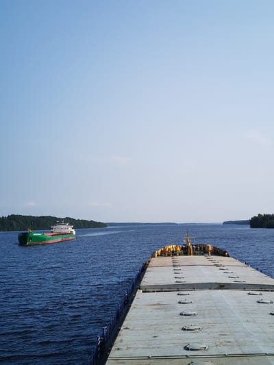 This media file depicts a wide, calm body of blue water, likely a river or lake, under a clear, pale blue sky. The scene is set with a bright, sunny day, suggesting fair weather.

In the distance, on the left side of the frame, a long green and red cargo ship is navigating the waterway. The ship has a white superstructure and appears to be moving forward, leaving a gentle wake. Its hull is primarily green, with a red stripe along the waterline. Text is visible on the side of the green hull, though it is too small to read.

In the foreground and dominating the right side of the image is the deck of another vessel, presumably the one from which the photo was taken. This vessel is a barge or a similar flat-topped cargo ship, with a large, grey, flat deck covered in numerous circular hatches. The deck has a weathered appearance. At the stern of this foreground vessel, a yellow structure with some poles and what appears to be a mast is visible.

The background is lined with lush green trees, forming a dense forest along the distant shorelines, suggesting a rural or natural setting. The overall impression is one of peaceful navigation on a calm waterway under a clear sky. The time of day appears to be daytime, likely mid-morning or afternoon, given the brightness and the quality of light. No people are clearly visible in the image. The location context is Irma, Russia.