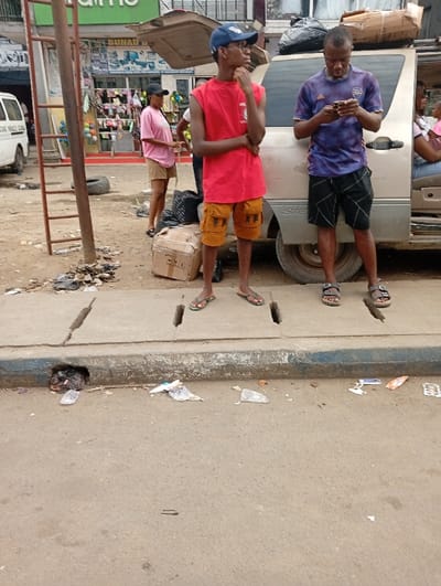 Three people are standing by a silver minivan with its trunk open. One man wears a blue hat and red sleeveless shirt, another a purple soccer jersey, looking at his phone. A woman in pink shorts stands nearby. A ladder is visible to the left, and the minivan's trunk is open. Garbage litters the gutter and street.