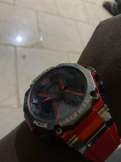 A close-up, slightly angled shot captures a person's left arm and hand, adorned with a distinctive red and silver G-Shock style watch. The watch features a robust silver bezel with visible text engravings: "START" on the left, "RESET" at the top, "SHOCK RESIST" on the upper right, "MODE" on the lower right, "LIGHT" at the bottom, and "G-SHOCK" on the lower left. The watch face is dark, with prominent red accents forming a geometric, almost propeller-like design in the center. While the watch appears to be an analog-digital model, the digital display is not clearly visible or illuminated. The watch strap is a vibrant red, contrasting with the silver bezel.
The arm, exhibiting a dark skin tone, is positioned with the watch prominent. The background reveals an indoor setting with light-colored, possibly beige or off-white, tiled flooring. A bright light source creates a noticeable glare on the reflective surface of the tiles, suggesting artificial illumination, which could imply evening or night, or simply an indoor environment in Shen, Nigeria with limited natural light. The focus is primarily on the watch and the arm, with the background slightly out of focus. No specific activity is captured beyond the person wearing the watch.
