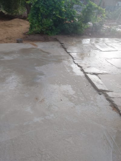 This is an outdoor, eye-level shot of a paved area that is wet, indicating it is raining or has recently rained. The paving stones are visible, with some cracks and unevenness between them. In the background, there is a patch of sandy soil and lush green foliage, suggesting a garden or yard. A small, grey bucket sits on the sand near the edge of the paved area. The scene is located in Kyzylorda, Kazakhstan.