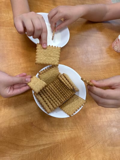 This image, located in the Gilboa Regional Council, Israel, presents an overhead view of four hands engaging with biscuits and liquid on a light brown wooden table surface. Two white ceramic bowls are visible. The upper bowl contains a white liquid, likely milk, into which a hand from the upper right is dipping a square, light brown biscuit. The lower bowl holds multiple square Osem Petit Beurre biscuits, some stacked vertically, others lying flat. A hand from the bottom left is in the process of picking up a biscuit from this bowl. Another hand from the top left holds a biscuit suspended above the lower bowl. A fourth hand, from the bottom right, holds a single biscuit. A small, partially visible decorative ceramic object with a floral pattern is present on the far right side of the wooden surface.