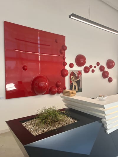 This image presents a modern interior scene of what appears to be a reception or office area located in Galatina, Italy, with no individuals present. The walls are uniformly white.
On the left wall, a large, rectangular, highly glossy red panel is mounted vertically. This panel features multiple embedded or affixed red spherical elements of various sizes, with one large sphere near the bottom-center and smaller ones predominantly along its right edge. Two subtle, horizontal indented lines are also present on the panel's surface. Adjacent to this, on the right wall section, a series of smaller, glossy red hemispherical forms are directly affixed to the wall in an arcing pattern. Below these, a framed portrait-oriented painting depicts a woman with vibrant red hair styled in an updo, wearing a dark top, with her right hand raised to her mouth holding what appears to be a lit cigarette or similar slender object, from which a wisp of smoke emanates.
A single, elongated, black linear LED light fixture is suspended horizontally from the white ceiling.
In the foreground, a white, multi-tiered, angular reception desk occupies the lower right portion of the image. Its design incorporates several horizontally stacked panels that appear to be internally illuminated. On the desk's top surface, a small white ceramic bowl containing light-colored, irregularly shaped objects is positioned next to a stack of rectangular, light-toned wooden blocks. A small white paper boat, inscribed with the text "Crescita, sempre e solo," also rests on the desk. Directly in front of the reception desk, a dark, angular planter box, likely black or dark gray, contains a bed of small, light-colored pebbles, from which a green, grass-like plant grows.
