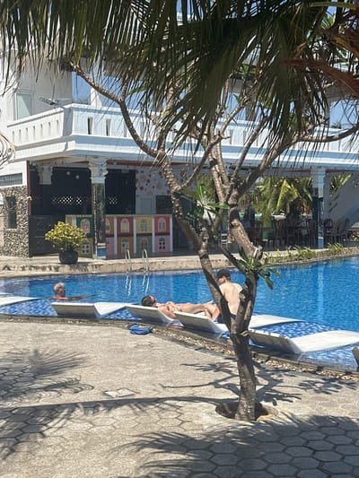 A poolside scene captures a sun-drenched day. Several individuals relax on lounge chairs beside a blue swimming pool. One person swims in the pool. Adjacent to the pool is a building with a painted facade reminiscent of colorful houses. A palm tree partially frames the view, casting shadows on the paved area surrounding the pool.