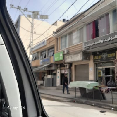 The image, captured from inside a vehicle, shows an urban street scene in Nuseirat, Palestinian Territories. The view is slightly angled upwards, revealing buildings with storefronts. Signage in Arabic is visible on several of the buildings. On the right side of the frame, an individual wearing dark clothing is walking down the sidewalk. Street furniture and merchandise are placed along the storefronts. A utility pole with wires is visible on the left side of the frame against a light sky. The vehicle's interior, including part of the dashboard and the edge of the car door, are visible in the foreground. A watermark "Galaxy A14" is present in the lower right corner.