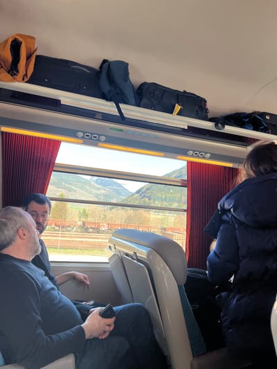 The scene is inside a train carriage. Two men and a woman are seated, facing the window. The woman is seated on the right side of the frame, facing the window. The man on the left side of the frame is seated and looking towards the window, while the other man is seated and looking to the right. All three people are wearing jackets. There are a few bags on the overhead rack. The window shows a view of a mountain range with green trees and a valley with a road and a river. The train is moving towards the mountains.