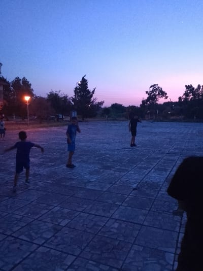 The image depicts a twilight scene in Gorno Aleksandrovo, Bulgaria. The sky is a gradient of deep blue at the top transitioning to pink and purple near the horizon, suggesting either dawn or dusk. In the foreground, a paved area with a checkerboard pattern of square tiles is visible. Several individuals are present in the scene. On the left, a young boy wearing a dark blue t-shirt and shorts is standing with his arms outstretched as if in motion. Further back, another boy in a blue t-shirt and shorts is standing, appearing to be talking on a phone or holding something to his ear. In the middle ground, a third boy, dressed in dark clothing, stands with one hand raised to his head. In the background, silhouetted trees and a street lamp with an orange glow are visible. The overall atmosphere is casual, possibly indicating recreational activity.