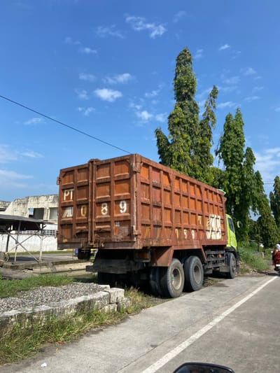A large, rusty dump truck is parked on the side of a paved road in Kecamatan Tulangan, Indonesia, at coordinates -7.477 latitude and 112.639 longitude. The truck is facing the right, with its bed tilted up. The truck is partially obscured by a row of bushes on the right side of the image, and tall, green trees on the left. The truck has a white, painted number "PT 089" on its side. It appears to have recently been used, with dust and dirt on the sides. The road is lined with a white strip on the right side of the image, and there are a couple of large concrete blocks by the truck. To the left of the truck, a row of bushes and a wall and gate can be seen in the distance. The sky is blue, with a few white clouds.  There is another, smaller vehicle driving away from the truck on the road in the distance. The truck's back tires are touching the road. The truck's front wheels are visible, but appear to be off the road.