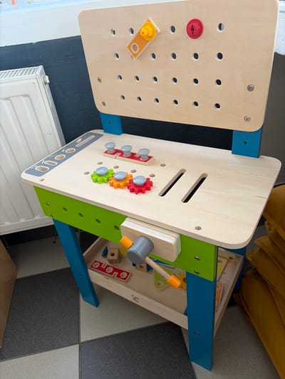 The image shows a wooden toy workbench with colorful accents. The workbench is made of light-colored wood and features various interactive elements designed for children. These include a pegboard with holes, several colorful gears (red, orange, and green), toy screws and bolts, and a toy hammer with a grey head and an orange handle. Some of the toy screws and bolts have red, yellow, and grey caps. The workbench is supported by blue and green wooden frames.
In the background, a white radiator is visible against a dark blue wall. The floor is a tiled pattern of light grey and black squares. The scene appears to be indoors, likely in a child's play area or playroom. The lighting suggests it is daytime. There is no visible text in the image. The overall impression is that of a well-equipped and engaging toy for children.