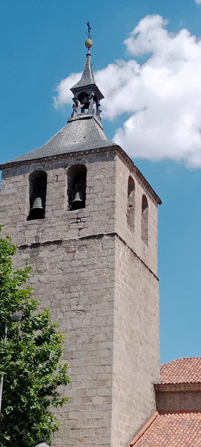 The image shows a stone bell tower in Galapagar, Spain, on a bright, partly cloudy day. The tower is square and constructed from rough-hewn stone blocks. It features two arched openings on its visible side, each containing a large bell. The tower is topped with a grey, multi-sided roof, which tapers to a point crowned with a weather vane shaped like a cross. Part of a red-tiled roof and a green tree are visible in the foreground, obscuring the base of the tower. The sky is a vivid blue with scattered white clouds.
