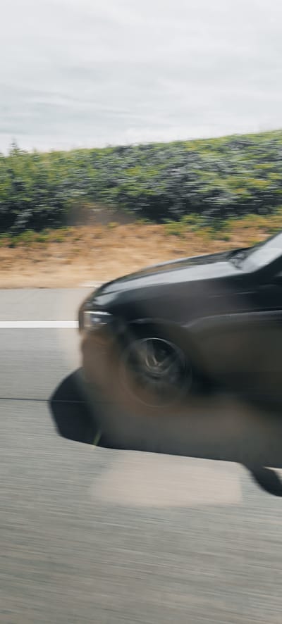 The image shows a blurry, motion-streaked view of a dark-colored car moving along a road in Waiblingen, Germany. The car's front end and a wheel are visible, with the rest of the vehicle blurred due to the speed. The road appears to be a highway with a white lane marker. To the right of the road, there is a blurred embankment covered in green vegetation and dry, brownish vegetation. The sky above is overcast with light clouds. The motion blur suggests the photograph was taken from another moving vehicle.