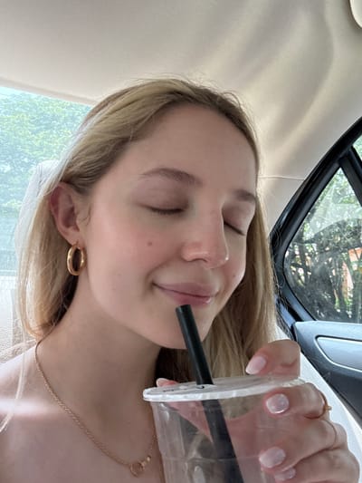 A young woman with blonde hair and closed eyes is drinking from a clear plastic cup with a black straw. She is wearing gold hoop earrings and a gold necklace with a circular pendant. Her fingernails are painted a light pink. The woman is sitting in the back seat of a car, with the interior of the car visible, including the headrest and window. Outside the car window, trees and foliage are visible, suggesting a location with greenery. The lighting is natural and appears to be daylight. The context of this scene is a woman enjoying a drink in a car in Coral Gables, United States of America.