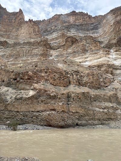 The image shows a wide, muddy river flowing in front of a massive, layered cliff face. The cliff is composed of various shades of brown, grey, and white rock, with distinct horizontal striations indicating geological stratification. The terrain at the base of the cliff is rocky and barren, with a single small, scrubby tree visible on the left side of the riverbank. The sky above is overcast with patches of white clouds. This scene is characteristic of the landscape found near Stongde, India, a region known for its dramatic mountainous terrain and glacial rivers.