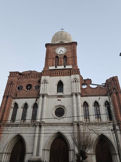 A low-angle, full-frame, eye-level shot captures the exterior of the San Fernando Cathedral in San Fernando, Chile. The cathedral, primarily constructed of red brick and white plaster, exhibits significant structural damage, with sections of the roof and walls appearing to be in disrepair. A large clock face is visible on the brick tower, indicating approximately 10:55. The sky is overcast and pale blue. The scene is devoid of human presence.