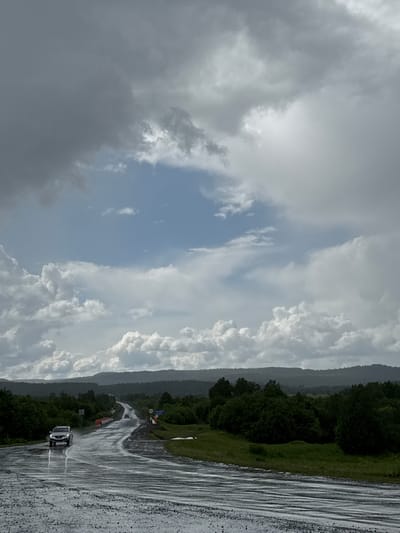 An overcast sky with dark, heavy clouds dominates the upper portion of the image, contrasting with patches of lighter, broken clouds and a sliver of blue sky. A two-lane road, wet from rain and reflecting the cloudy sky, curves through a landscape of rolling hills covered in dense green trees. The road appears to be wet and somewhat muddy in the foreground. Several vehicles are visible on the road. A silver SUV is in the left lane, moving away from the viewer, its headlights on. Further down the road, two other vehicles are visible, also appearing to be moving away. The roadside is lined with lush green vegetation and trees. In the distance, the hills are covered in a dark, uniform layer of trees, creating a textured horizon. The scene suggests a rural or remote location, possibly during or after a rain shower. The specific location of Kazakkulovo, Russia, is not visually evident from the image itself, but this description pertains to the visual elements present.