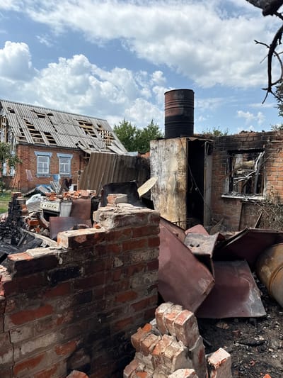 The image depicts a scene of severe structural damage in Nedryhailiv, Ukraine, under a partly cloudy sky. In the immediate foreground, a charred, partially collapsed brick wall stands. To its right, a dark, rusted, inverted metal drum lies on the ground amidst fragmented debris. Adjacent to the foreground wall, in the mid-ground, a severely burnt brick structure with a hollow, charred window frame and a damaged doorway is visible. Resting atop an adjacent smaller, singed, corrugated metal-sided structure is a vertical, rusted metal barrel. The ground surrounding these structures is covered with ash, charred rubble, and large, rusted, twisted metal sheets. To the mid-left, a two-story red brick house with white-framed windows exhibits extensive roof damage, with significant portions of its corrugated roof missing, exposing the underlying wooden rafters. Near the base of this house, piles of household debris, including a white kitchen range, are scattered. No individuals are present in the scene. Green foliage from trees is visible in the background.