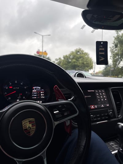 The image provides an inside view of a car, likely a Porsche, on a cloudy day in Wydminy, Poland. The focus is on the steering wheel and dashboard. A "VIP" tag hangs from the rearview mirror, suggesting a special occasion or status. The dashboard displays various information including vehicle speed (indicated by "GTS"), engine temperature (93°C and 99°C), and tire pressure (1.2 bar). The infotainment system is visible, showing a phone dial pad with "Mobile Vikings" and "CARIBI" text. The time displayed is 16:30, indicating late afternoon. Outside the car, a traffic light and trees are partially visible through the windshield, suggesting the car is stationary, possibly at a traffic light or in traffic. The overcast sky implies a likely chance of rain. The image is taken from the driver's perspective.