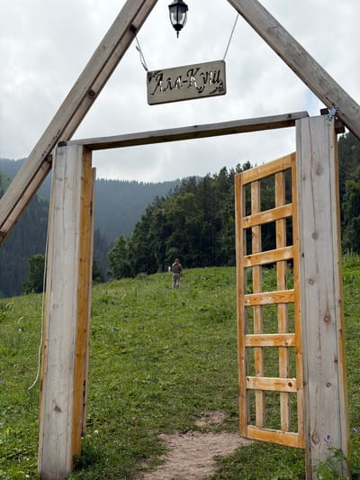 An outdoor, medium shot captures a wooden archway framing a lush green grassy hill. A person in the distance walks towards the right, facing away from the camera. The archway features a wooden gate with a lattice design on the right side, which is slightly ajar. Above the archway, a sign with Cyrillic lettering hangs from a chain. The background consists of a dense forest covering rolling hills, with a cloudy sky overhead. The scene appears to be in the Ala-Kuu valley near Kaindy, Kyrgyzstan.
