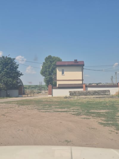 This is a daytime, eye-level, outdoor, full shot image. The scene depicts a rural or semi-rural area. In the foreground, there is a dirt road with tire tracks and patches of dry grass. To the left, a large tree with green foliage stands near a building with a curved, translucent roof, possibly a greenhouse or an industrial building. A brick and concrete wall runs along the edge of this structure.
Further into the scene, a light yellow building with a brown roof and brown trim is prominently featured. This building appears to be a two-story house with a small, rectangular window on the upper floor. A brick column is visible on the right side of this building. In front of the yellow building, there is a low white wall. Stacked concrete slabs are placed on the ground in front of this wall, atop a patch of overgrown grass.
In the background, a vast expanse of green fields stretches towards the horizon under a clear blue sky with scattered white clouds. Several electrical poles with wires are visible in the distance, along with a dark plume of smoke rising from behind a row of trees. The overall impression is one of a moderately developed rural landscape on a sunny day.
