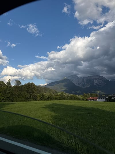 The image captures a scenic view of the Bavarian Alps in Bischofswiesen, Germany. In the foreground, a vibrant green meadow stretches out, with a wire fence running horizontally across it. Beyond the meadow, a dense line of dark green trees forms a natural barrier, behind which a majestic mountain range dominates the background. The mountains are rugged and imposing, with some appearing in shadow due to heavy, dark clouds overhead, while others are illuminated by sunlight breaking through. A few buildings with red-tiled roofs are nestled in the landscape at the base of the mountains. The sky is a striking contrast of deep blue and dramatic white and grey clouds, suggesting dynamic weather conditions.