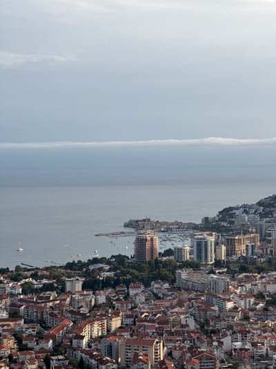 This is an aerial, long-shot, high-angle, eye-level, exterior, daytime, wide-angle, over-the-shoulder, vertical, 4K, RAW, shot of Markovići, Montenegro, overlooking the Adriatic Sea. The sky is a hazy blue with a distinct band of elongated clouds stretching horizontally across the mid-distance. The sea is calm and a muted blue-gray. In the distance, a town with red-tiled roofs and a small church steeple is visible on a peninsula jutting into the sea, with a marina full of boats. Beyond the town, modern high-rise buildings rise up along the coastline. The foreground is dominated by a dense urban landscape of residential buildings with red-tiled roofs and light-colored facades, interspersed with green trees. Several sailboats and motorboats are visible in the water. The overall impression is of a coastal city nestled between the sea and hilly terrain.