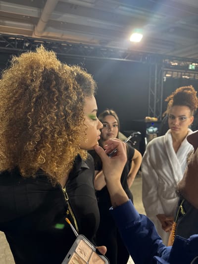 A backstage scene in a dimly lit, industrial-style environment, possibly a studio or event space in Issy-les-Moulineaux, France, depicts three individuals, with a makeup application in progress. In the foreground, a light-skinned female model with voluminous light brown curly hair wears a black collared top with a gold zipper. Her eyes are closed, revealing bright green eyeshadow on her eyelids. A partially visible individual, likely a male makeup artist with dark skin and a beard, wearing a blue long-sleeved shirt, is applying makeup to the model's chin area with a hand while holding a multi-shade makeup palette containing various light-toned powders/creams and a small brush with an orange tip.

In the midground, to the left of the central model, a female individual with dark hair, wearing a black top, observes the process, holding a thin, silver-colored tool. To the right of the central model, another light-skinned female individual, presumably a model, with dark curly hair styled upwards, wears a white bathrobe and has visible blue eyeshadow. She is looking towards the central model. The background features dark metal truss structures and overhead bright artificial lighting fixtures. The floor appears to be a dark, textured surface.