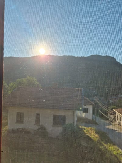 The image captures a view of the city of Lesidren, Bulgaria, through a window screen. The sun is setting behind a mountain, casting a warm glow on the scene. In the foreground, a house with a red tiled roof is visible, with two windows and a door. The house appears to be a typical residential building in the area. Behind the house, other buildings and a road can be seen, suggesting a village or small town setting. The natural environment is characterized by rolling hills covered in greenery. The overall impression is one of a peaceful rural landscape.