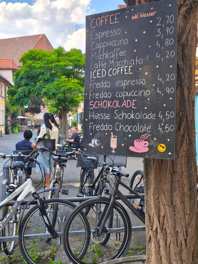 The image depicts a street scene in Erlangen, Germany, with a prominent chalk menu board attached to a tree. The menu lists various coffee drinks with their prices. In the foreground, several bicycles are parked. In the background, people are visible, including a woman standing with a purse, a man sitting on a bench, and another man in a motorized wheelchair. Buildings and lush green trees line the cobblestone street, suggesting a pleasant outdoor setting. The sky is partly cloudy. The menu board lists items like "Espresso", "Cappuccino", "Milchkaffee", "Latte Macchiato", and "ICED COFFEE" with prices in Euros. Below these, it lists "Freddo espresso", "Freddo capuccino", "SCHOKOLADE" (Chocolate), "Heisse Schokolade", and "Freddo Chocolate" with their respective prices. The menu board also features small decorative drawings of a drink and a coffee cup, as well as a smiley face sticker with hearts. The overall atmosphere appears to be that of a casual cafe or street vendor operating in a European city.