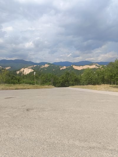 The image depicts a scenic landscape in Rozhen, Bulgaria. In the foreground, there is an asphalt road, suggesting a viewpoint or a park area. Beyond the road, a lush green forest extends, with dry grass and vegetation in the transition zone. The main focus of the background is a dramatic geological formation of sand and clay pyramids, characteristic of the region, against a backdrop of rolling green mountains. The sky is filled with dramatic, heavy clouds, indicating either approaching rain or overcast weather. The lighting suggests it is daytime, likely late afternoon given the dramatic cloud cover and muted colors. There are no people visible in the image. A wooden post with two small signs is present near the edge of the road in the mid-ground, but any text on them is illegible. The overall mood is one of natural grandeur and perhaps a touch of dramatic weather.